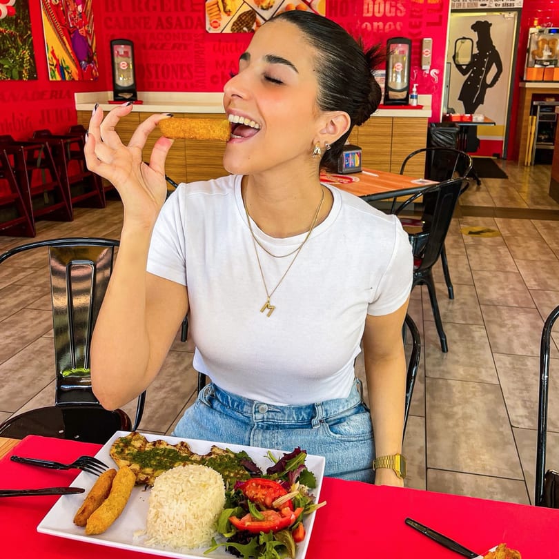 A woman in a white shirt and jeans smiles, holding a fried food stick to her mouth at La Perrada.