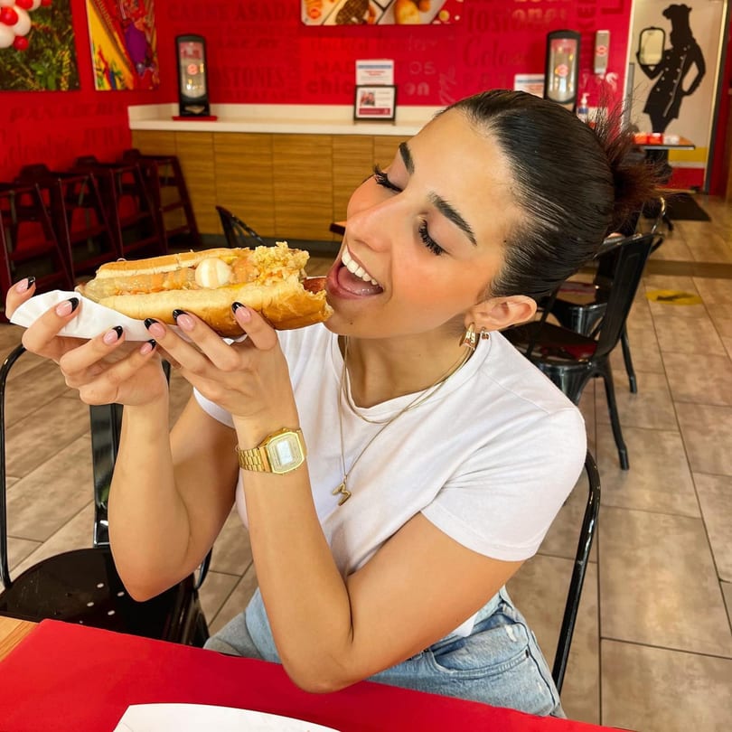 A woman in a white shirt and jeans happily takes a bite of a loaded hot dog at La Perrada.