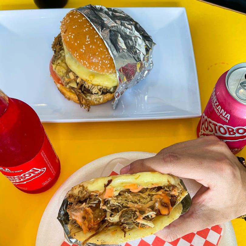 A La Perrada burger, an arepa, and two Postobón sodas on a yellow table.