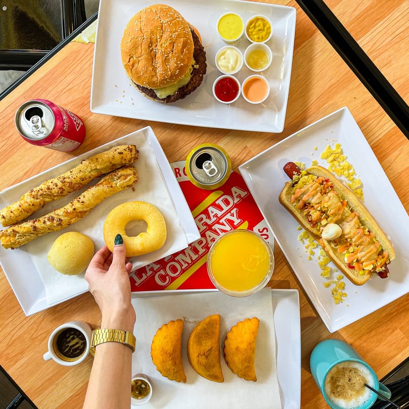 Overhead view of a table laden with a burger, hot dog, pastries, empanadas, drinks, and sauces.