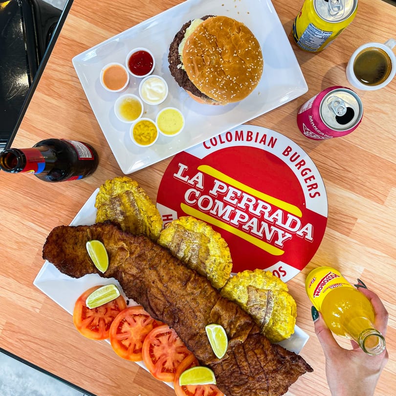 A La Perrada Company burger, fried fish, plantains, sauces, and drinks on a wooden table.