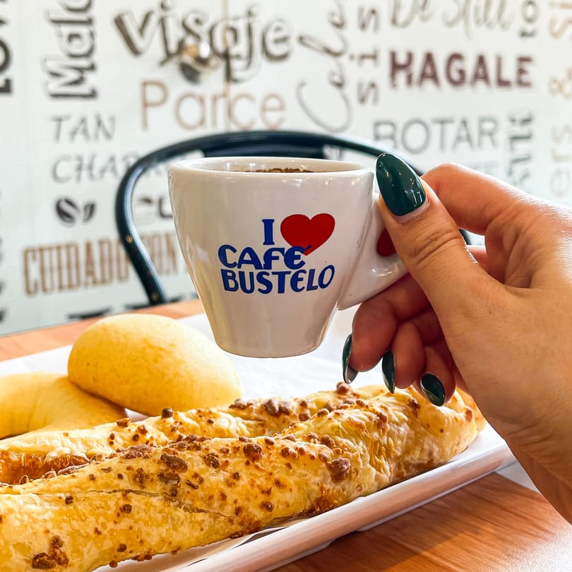 Hand holding a white cup of "I love Cafe Bustelo" coffee over cheesy breadsticks and rolls.