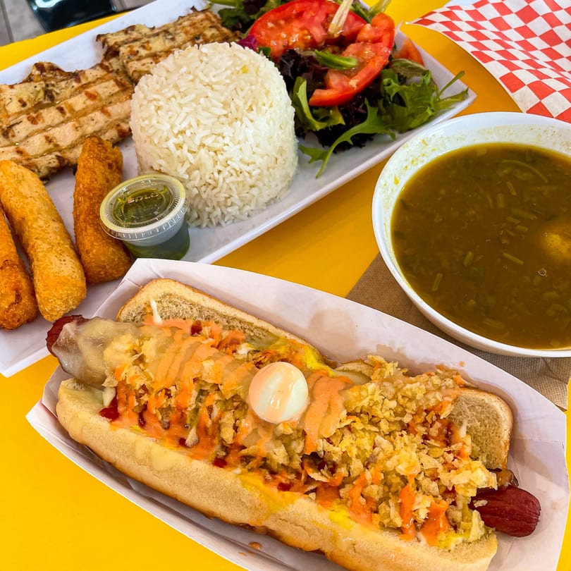 A La Perrada hot dog with toppings, a plate of grilled chicken, rice, salad, and a bowl of soup.