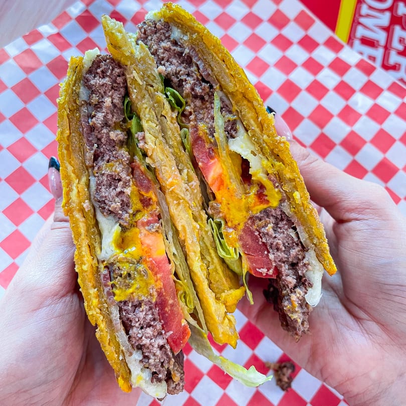 Hands holding a La Perrada burger, cut in half, revealing beef patties, cheese, lettuce, and tomato.